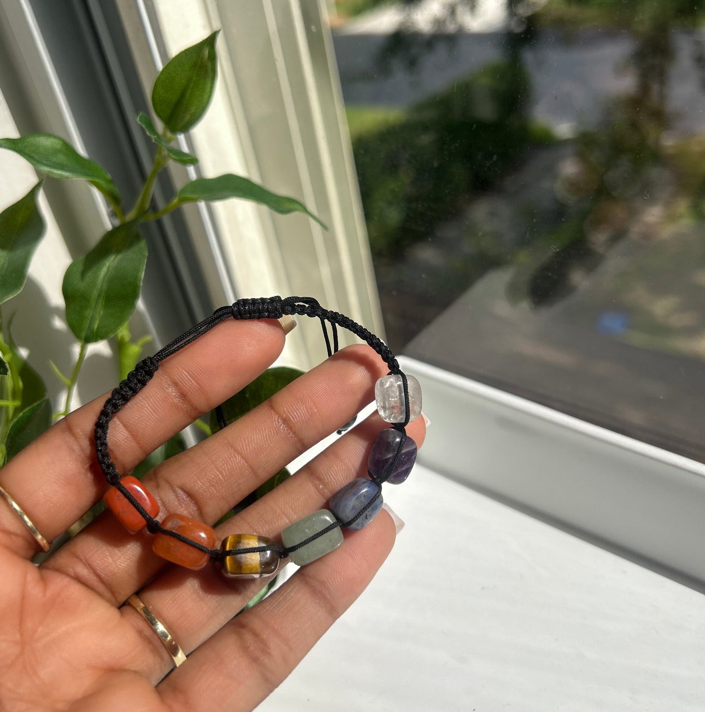 A hand holding a black cord bracelet with polished gemstone beads in purple, clear, orange, and yellow, set against a window background with green leaves.