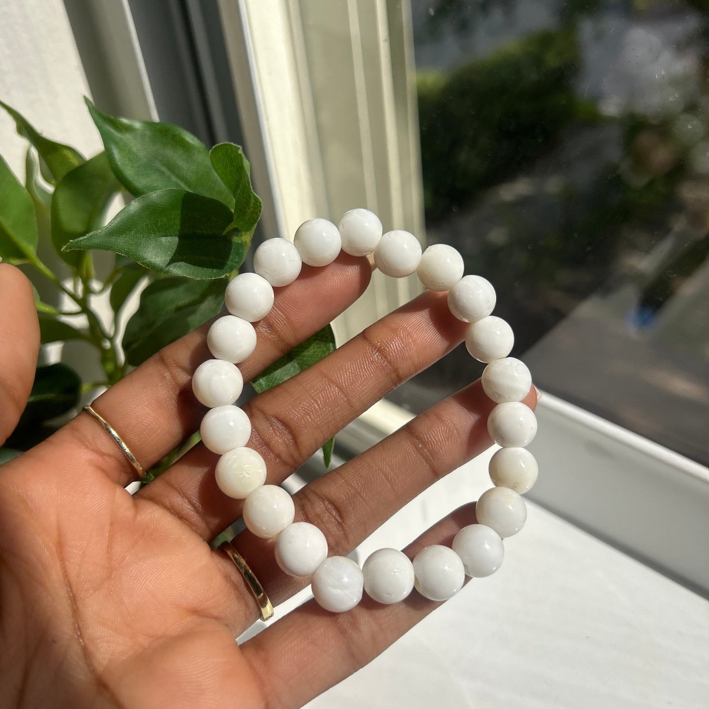 A white beaded bracelet held in a person's hand near a window with green leaves in the foreground.