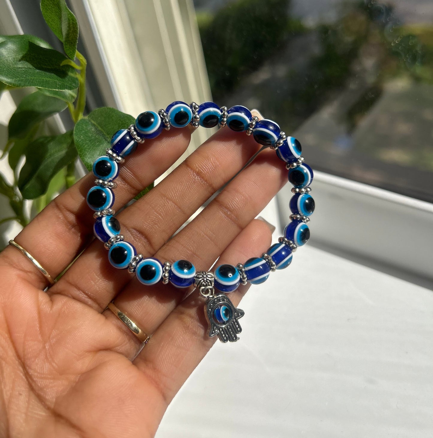 A blue and white evil eye beaded bracelet with a silver hamsa hand charm, held in a person's hand near a window.