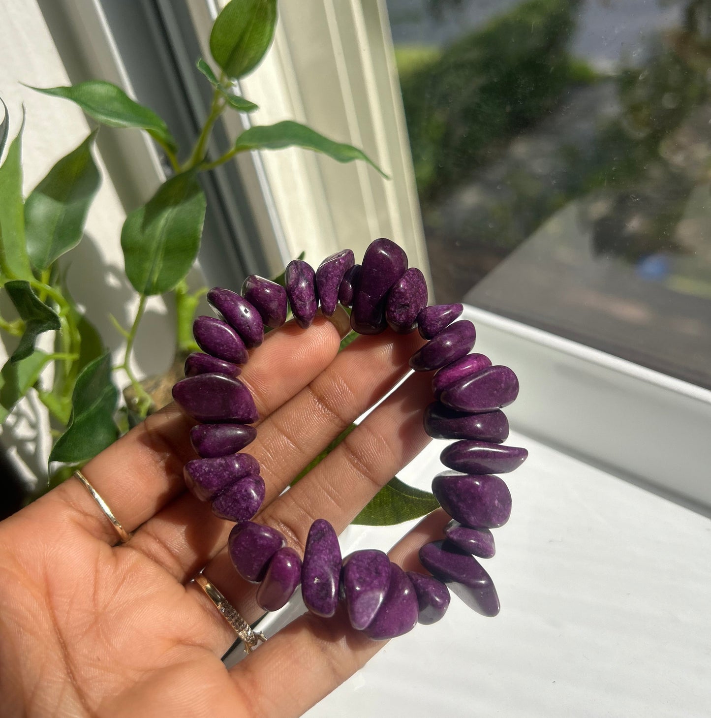 A purple tumbled stone beaded bracelet held in a person's hand near a window with green leaves in the foreground.