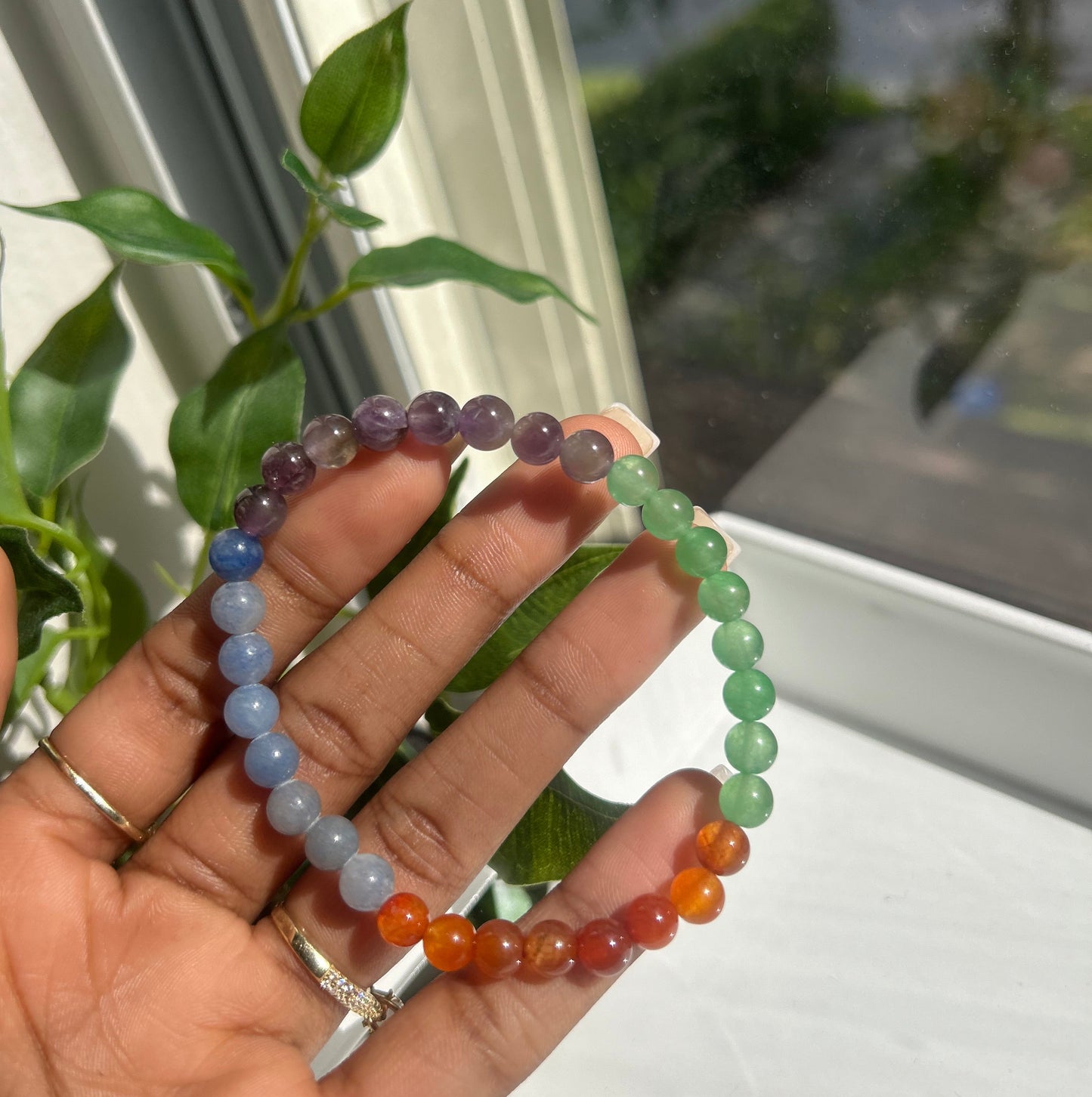A person's hand holding two beaded stretch bracelets. One bracelet features green beads, and the other features orange, red, purple, and blue beads, set against a background of a window and green leaves.