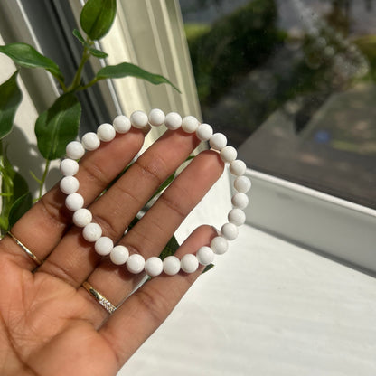 A white beaded bracelet held in a person's hand, with a window, green leaves, and a white wall in the background.