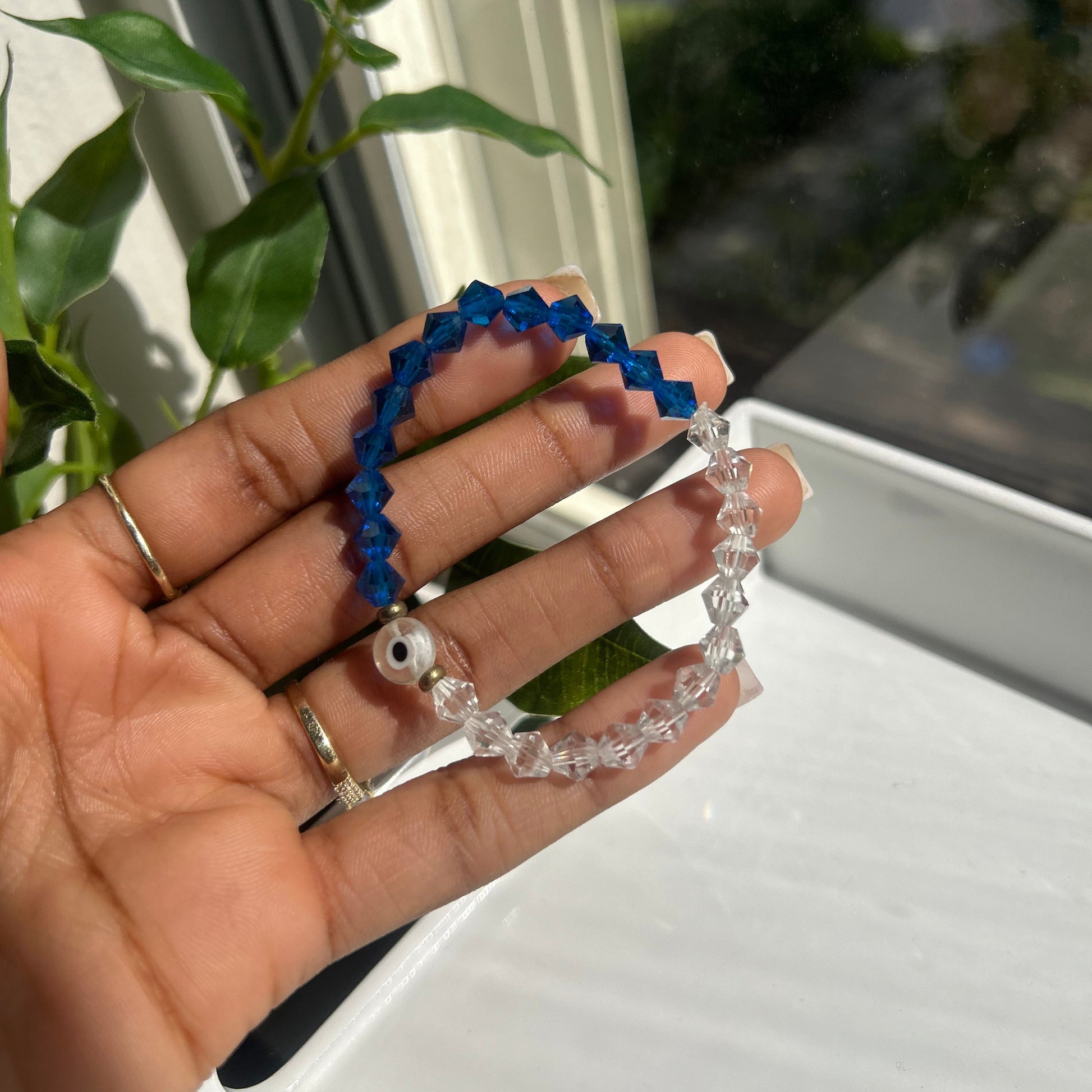 A hand holding two beaded bracelets, one made of dark blue beads and one made of clear faceted beads with a small white evil eye charm, in front of a window.