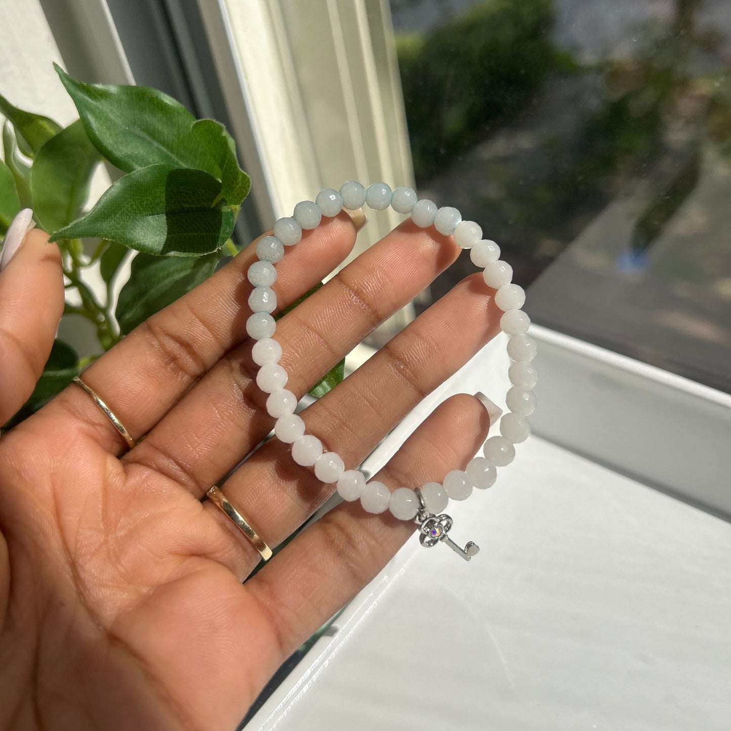A beaded bracelet made of small white oval beads with a silver clasp, held in a person's hand near a window with green leaves in the foreground.