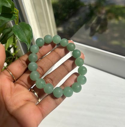 A beaded stretch bracelet made of light green jade-like round beads, held in a person's hand near a window with green leaves visible in the foreground.