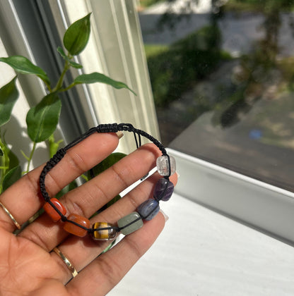 A hand holding a black cord bracelet with polished gemstone beads in purple, clear, orange, and yellow, set against a window background with green leaves.