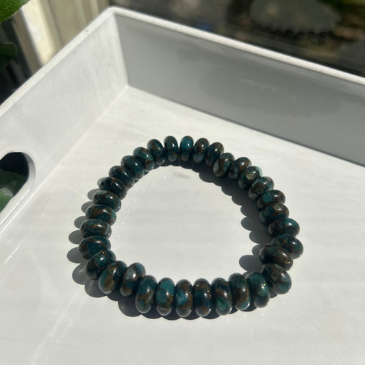 A beaded stretch bracelet made of dark, polished oval beads with a deep teal-blue and black marbled pattern, resting on a white surface.