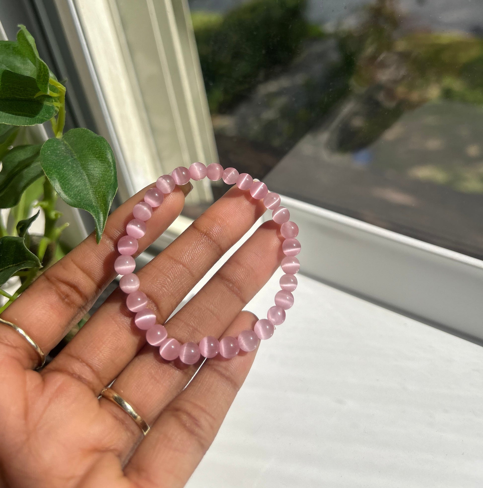 A pink beaded stretch bracelet held in a person's hand near a window, with green leaves and a bright outdoor view in the background.
