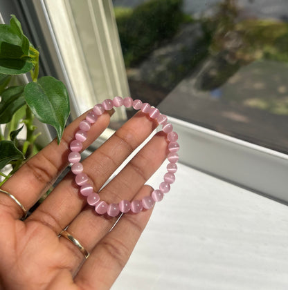A pink beaded stretch bracelet held in a person's hand near a window, with green leaves and a bright outdoor view in the background.
