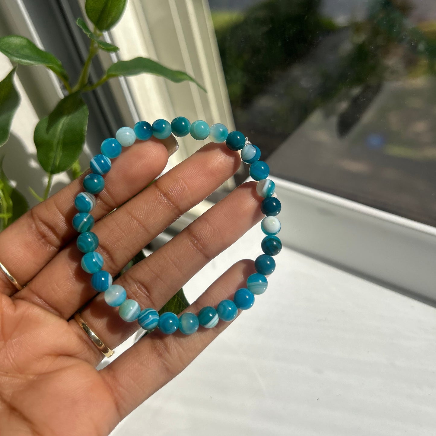 A beaded stretch bracelet made of round turquoise-blue and white marbled beads, held in a person's hand near a window with green leaves visible in the background.