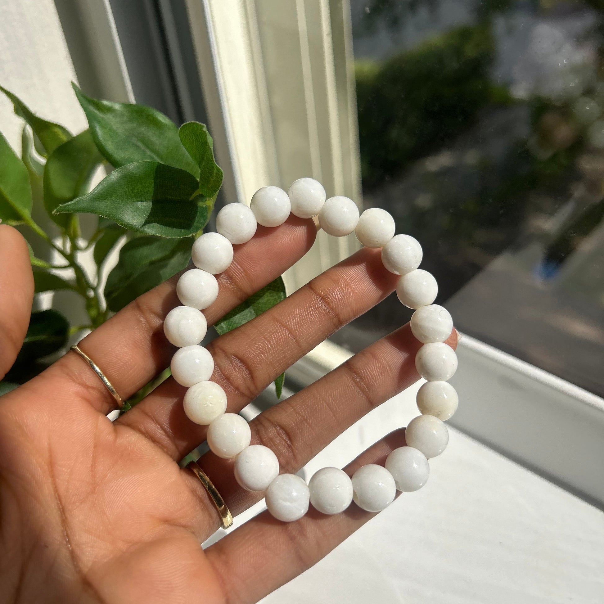 A white beaded bracelet held in a person's hand near a window with green leaves in the foreground.