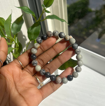 A beaded stretch bracelet made of polished gray and white marbled gemstone beads, held in a person's hand near a window with green leaves in the foreground.