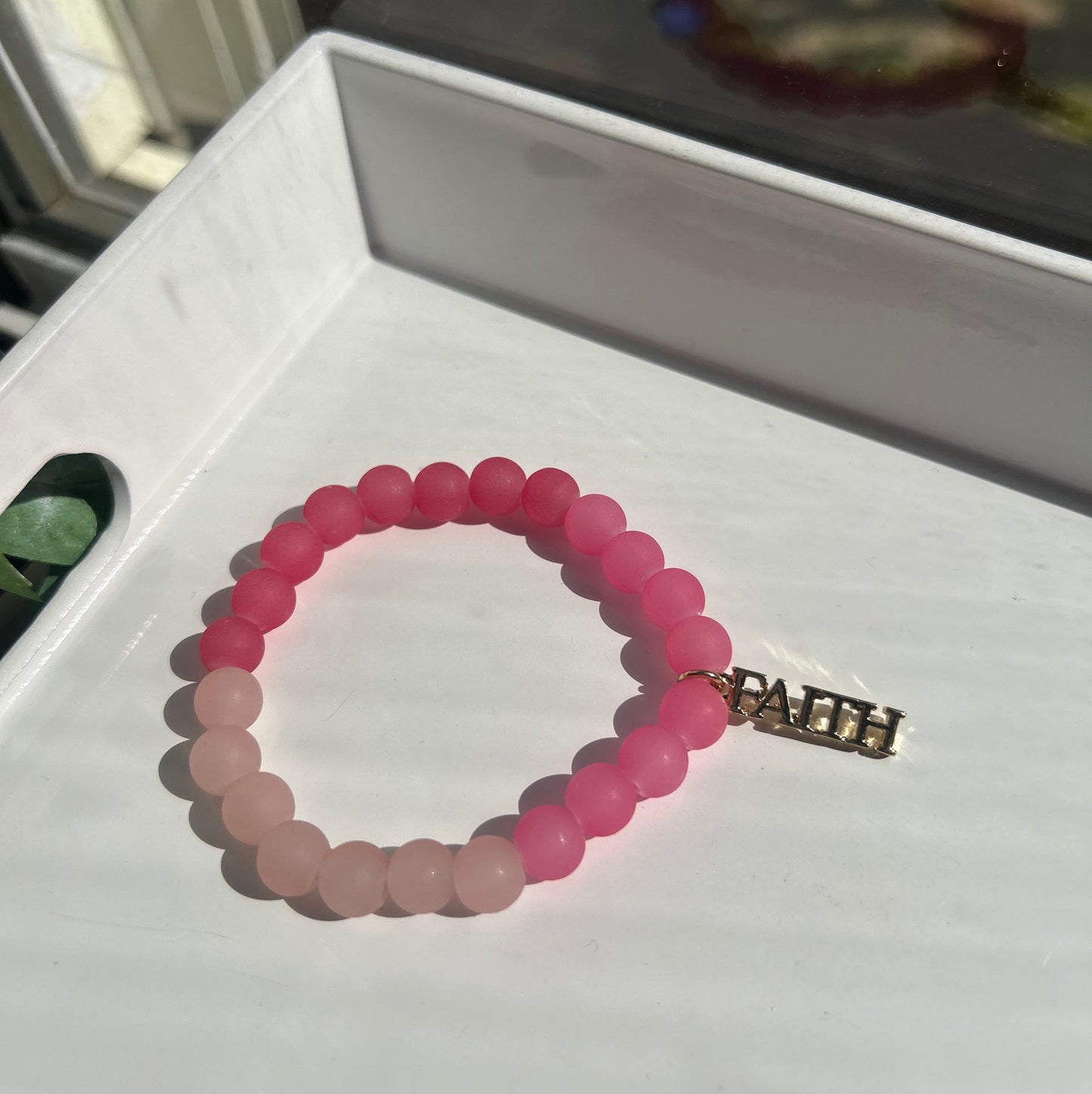 A beaded stretch bracelet with a gradient of pink beads transitioning from a darker magenta to a lighter blush pink, featuring a gold-toned metal clasp, displayed on a white surface.