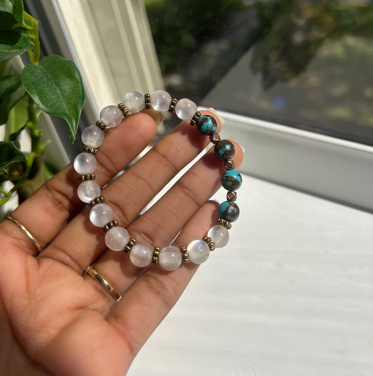 A hand holding a beaded stretch bracelet with white moonstone-like round beads, turquoise blue beads, and small bronze spacer beads, photographed near a window.