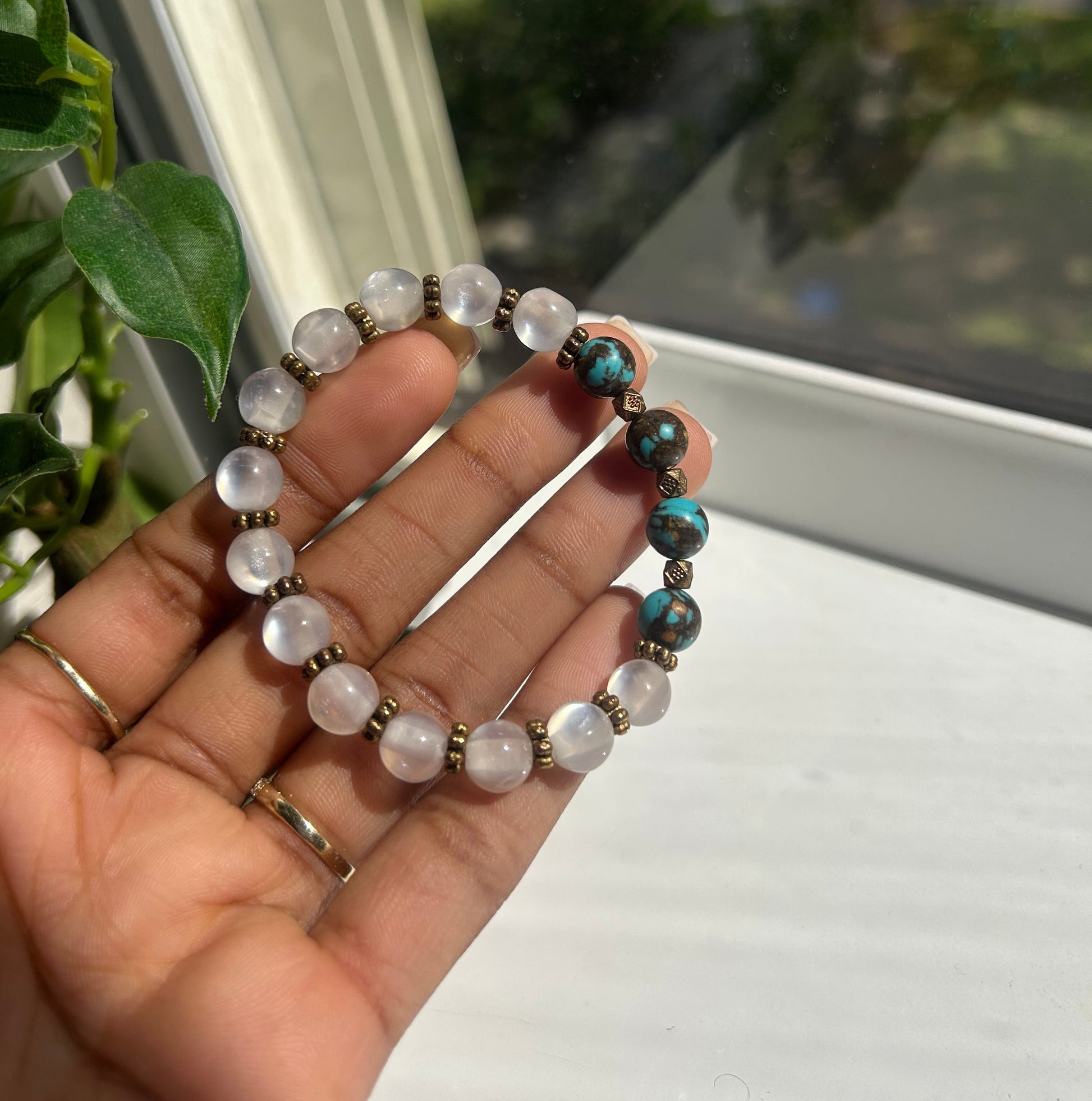 A hand holding a beaded stretch bracelet with white moonstone-like round beads, turquoise blue beads, and small bronze spacer beads, photographed near a window.