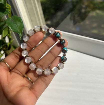 A hand holding a beaded stretch bracelet with white moonstone-like round beads, turquoise blue beads, and small bronze spacer beads, photographed near a window.
