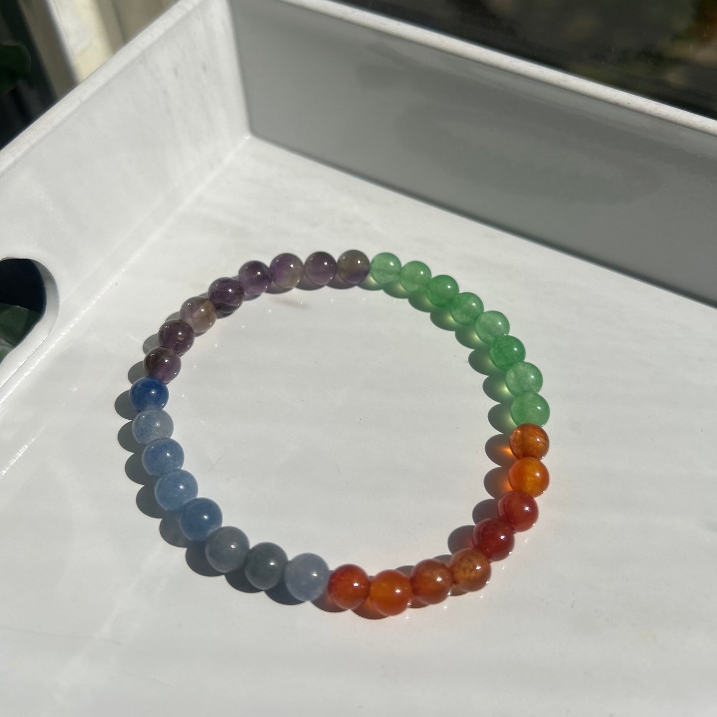 A beaded stretch bracelet arranged in a rainbow sequence of translucent gemstone-like beads, resting on a white surface.