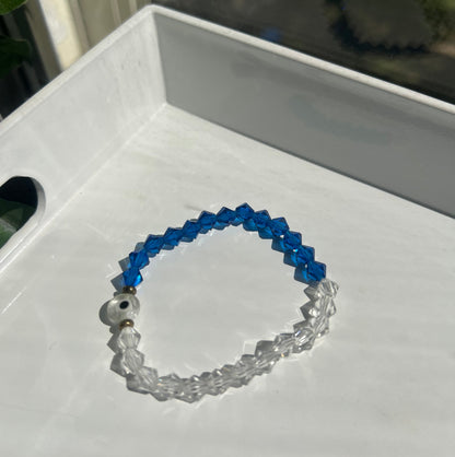 A beaded stretch bracelet with blue faceted beads on one side and clear faceted beads on the other, featuring a small white bead with a black center, resting on a white tray.
