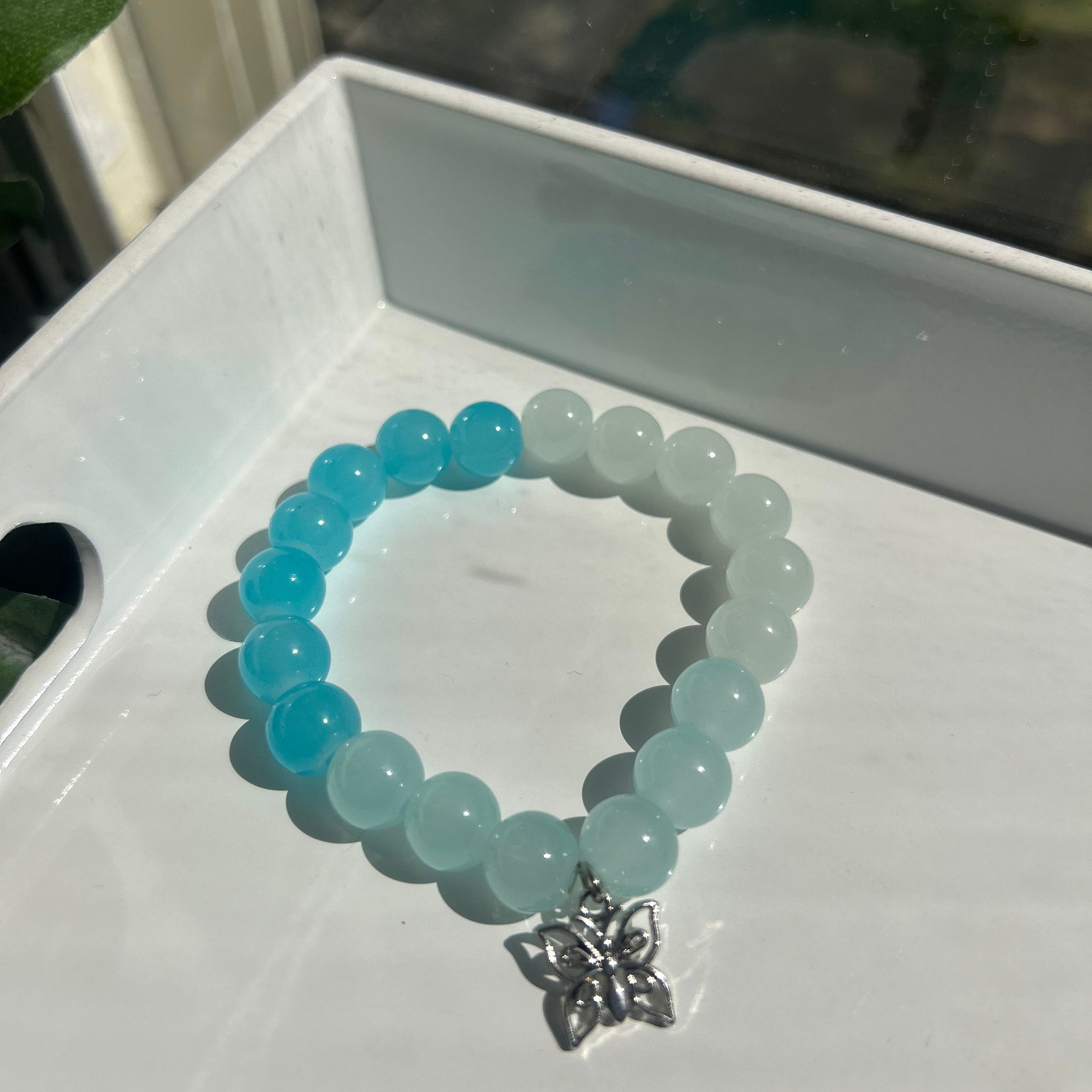 A beaded stretch bracelet with a gradient of light aqua to sky blue translucent beads, featuring a small silver butterfly charm, displayed on a white tray.