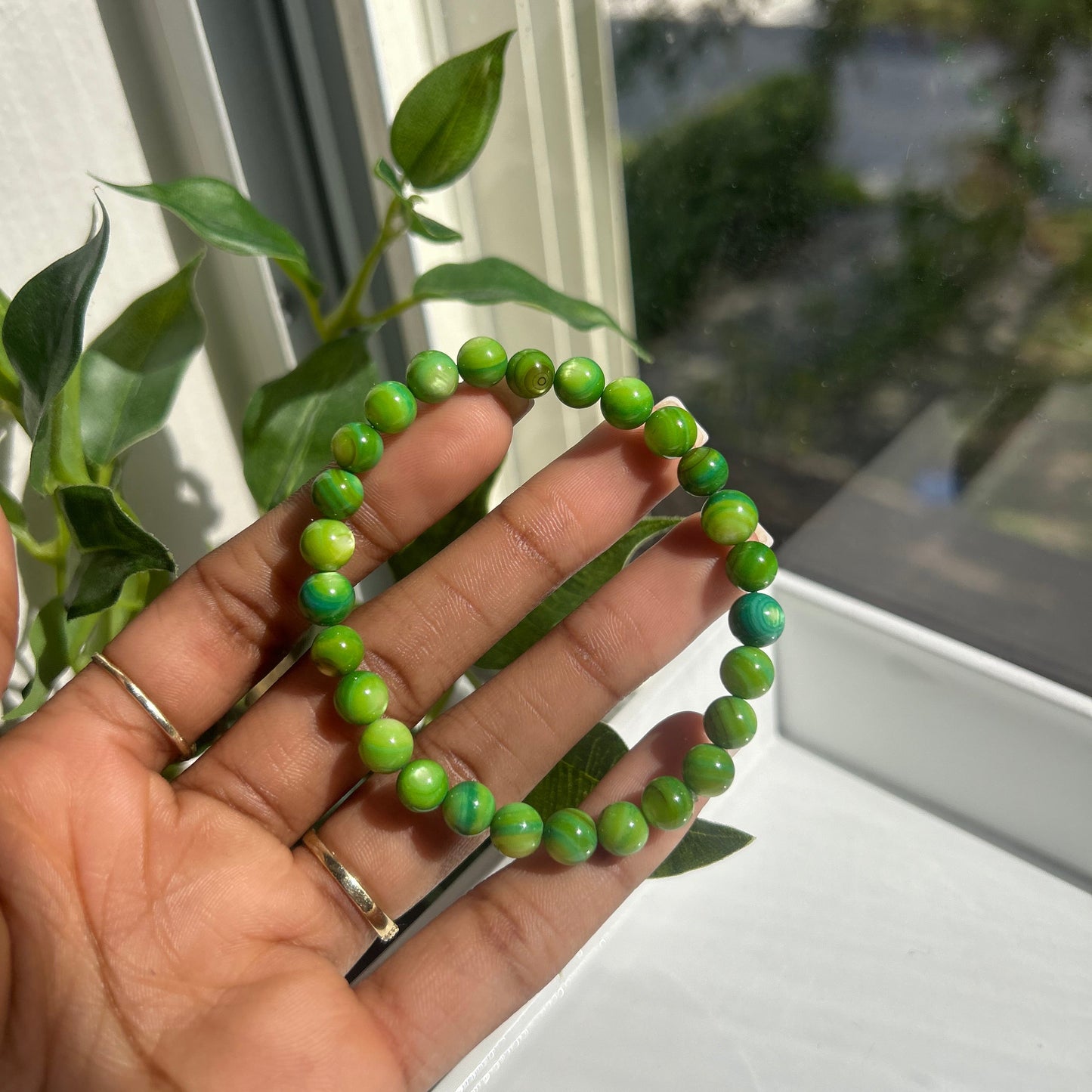 A green beaded bracelet held in a person's hand, with a window and green leaves visible in the background.