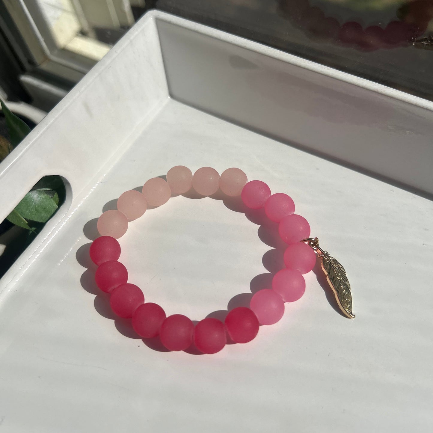 A beaded stretch bracelet with a color gradient transitioning from pale pink to vibrant magenta, featuring a small gold-toned leaf charm at the top, displayed on a white tray.