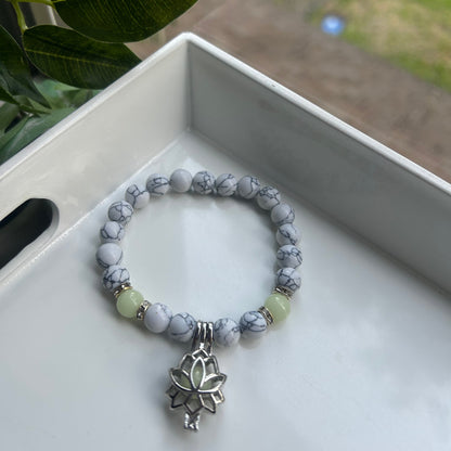 A beaded stretch bracelet with gray and white marbled beads, a light green bead, and a silver-toned lotus charm, displayed inside a white tray.