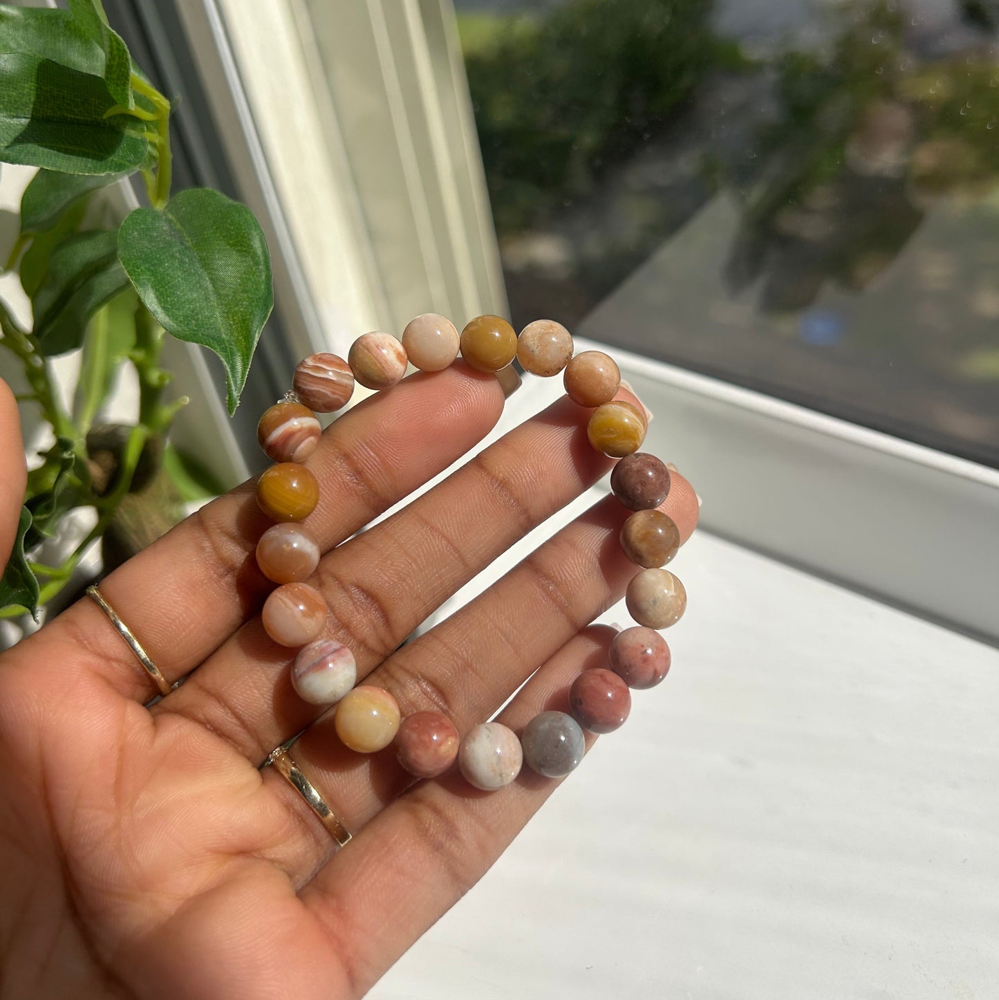 A beaded bracelet made of round, polished stone beads in earthy brown, beige, pink, and white tones, held in a person's hand near a window.