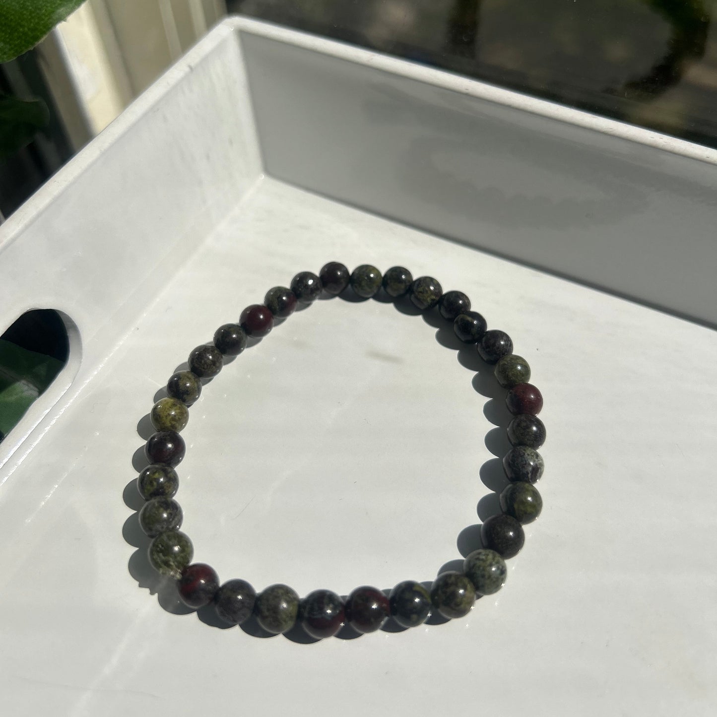 A beaded stretch bracelet made of dark, polished round beads with subtle green and reddish-brown tones, resting on a white tray.