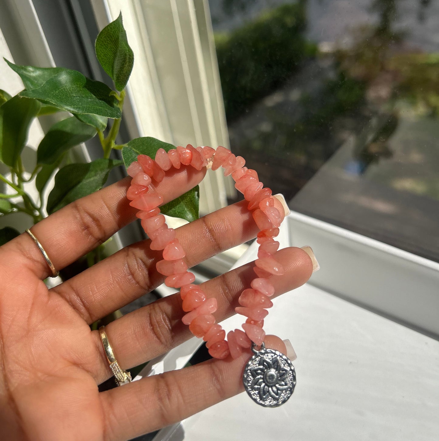 A person's hand holding a peach-pink chunky stone bracelet with a silver-toned circular charm, set against a window background with green leaves.