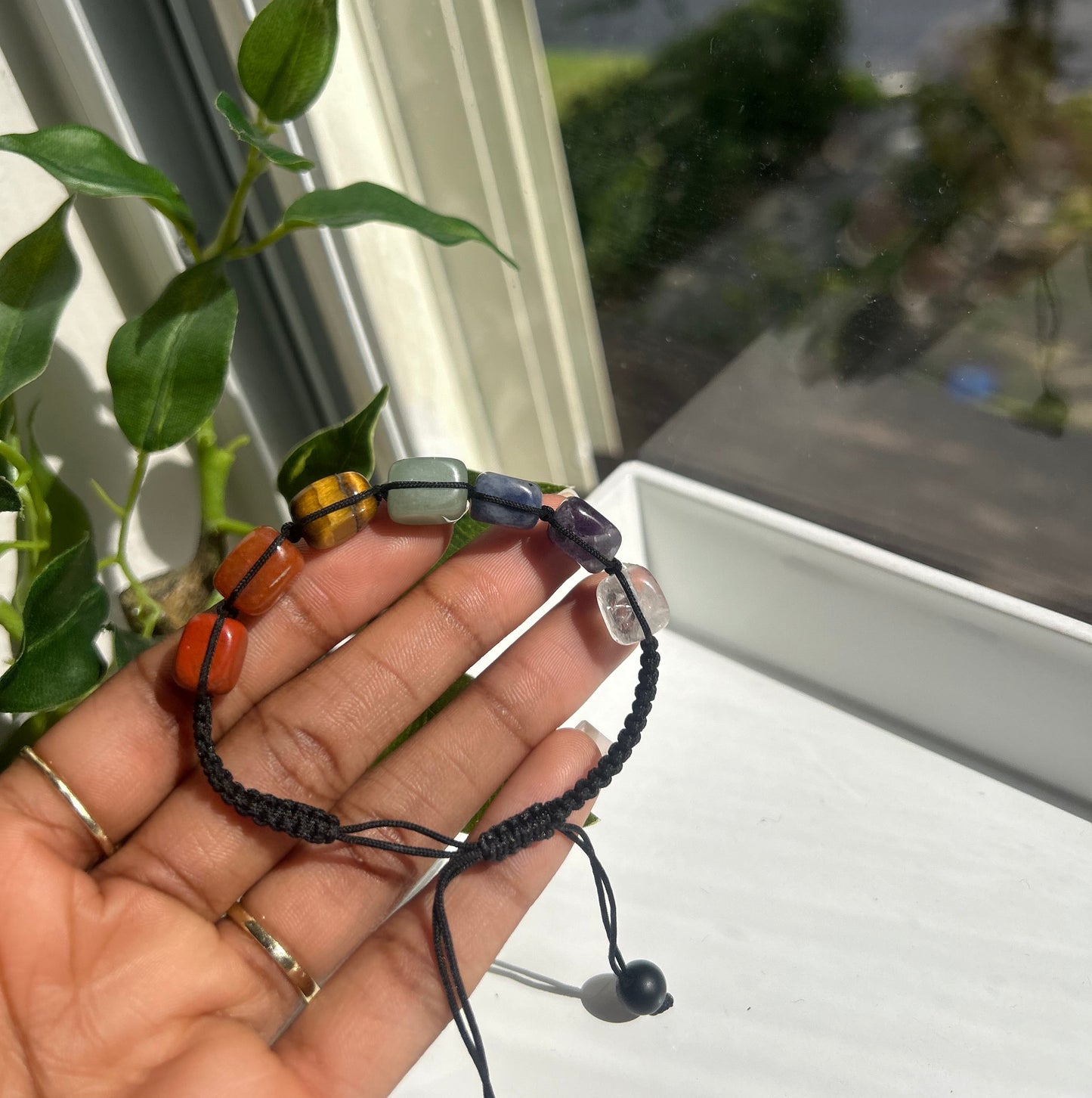 A person's hand holding a beaded cord bracelet with colorful round beads and a small gold-toned bell, photographed near a window with green leaves in the foreground.