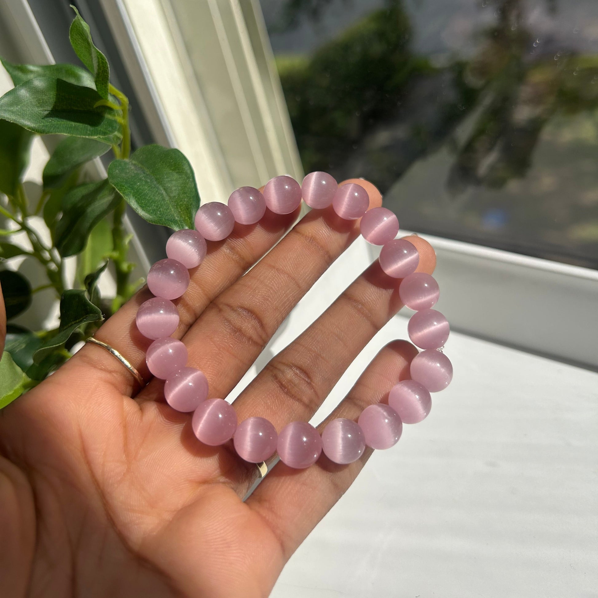 A person's hand holding a pink cat-eye bead bracelet in front of a window with green plants visible outside.