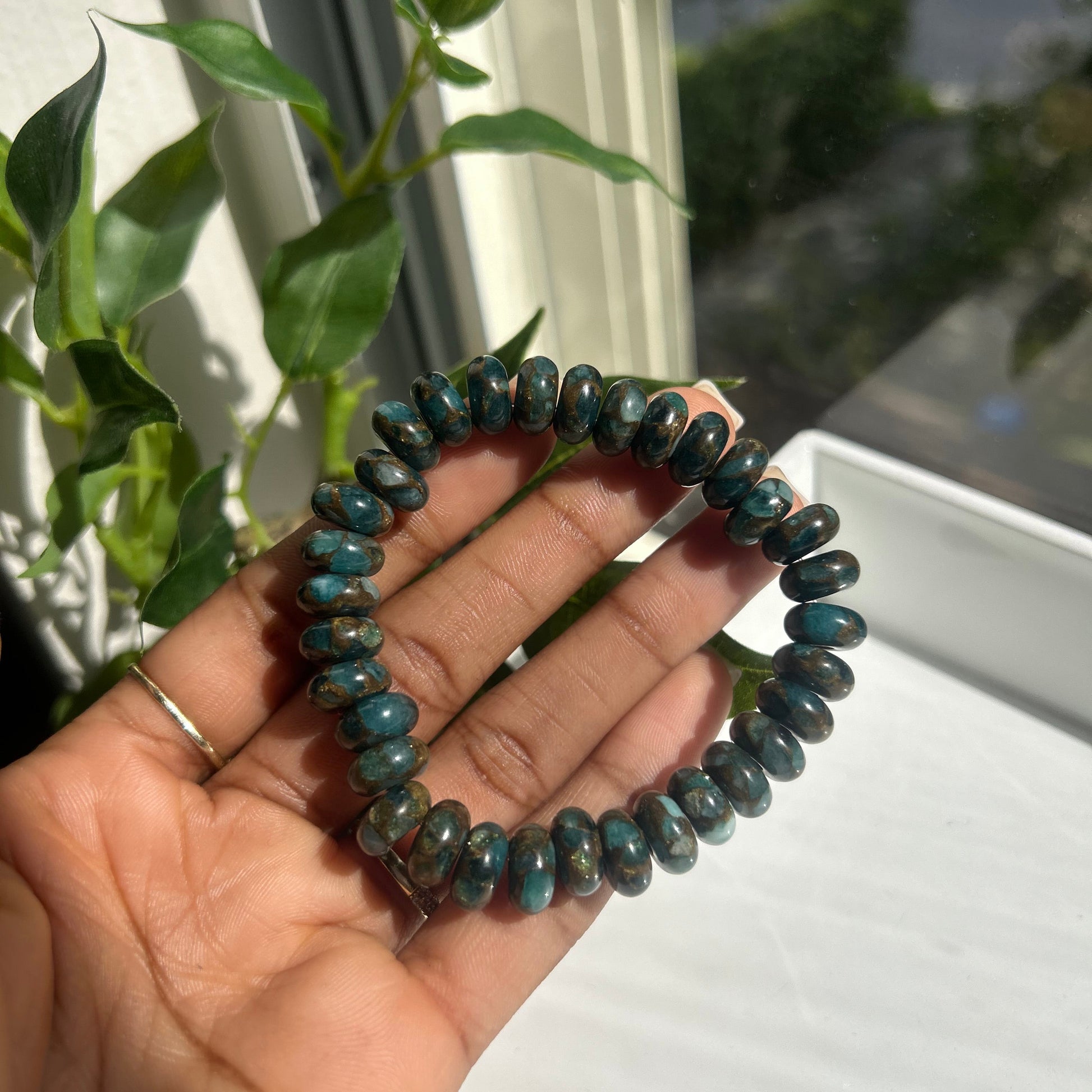 A beaded stretch bracelet made of polished dark teal-blue oval beads with metallic gold edges, held in a person's hand near a window with green leaves in the background.