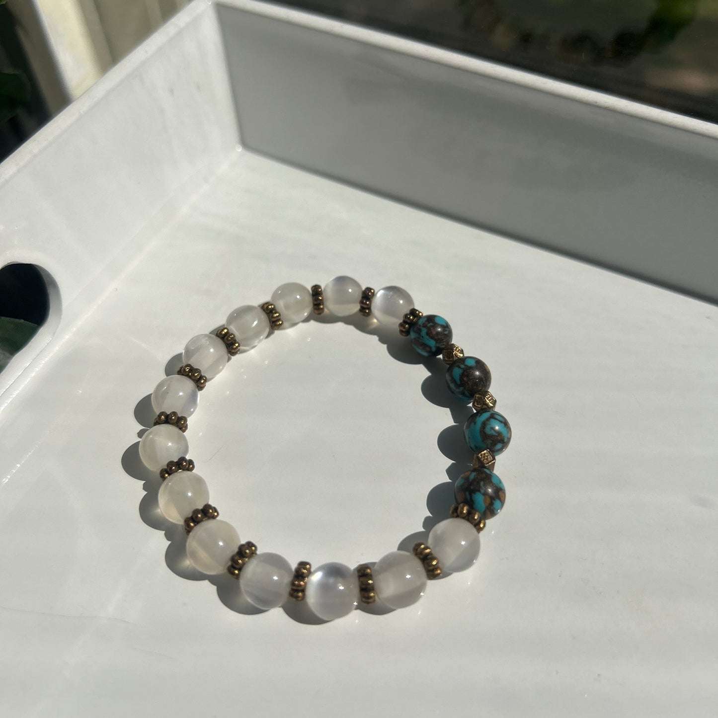 A beaded stretch bracelet with white translucent round beads, turquoise blue marbled beads, and small bronze spacer beads, displayed on a white surface.