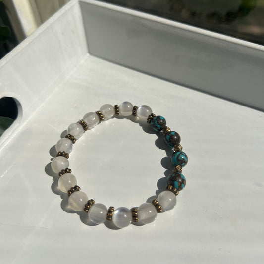 A beaded stretch bracelet with white translucent round beads, turquoise blue marbled beads, and small bronze spacer beads, displayed on a white surface.