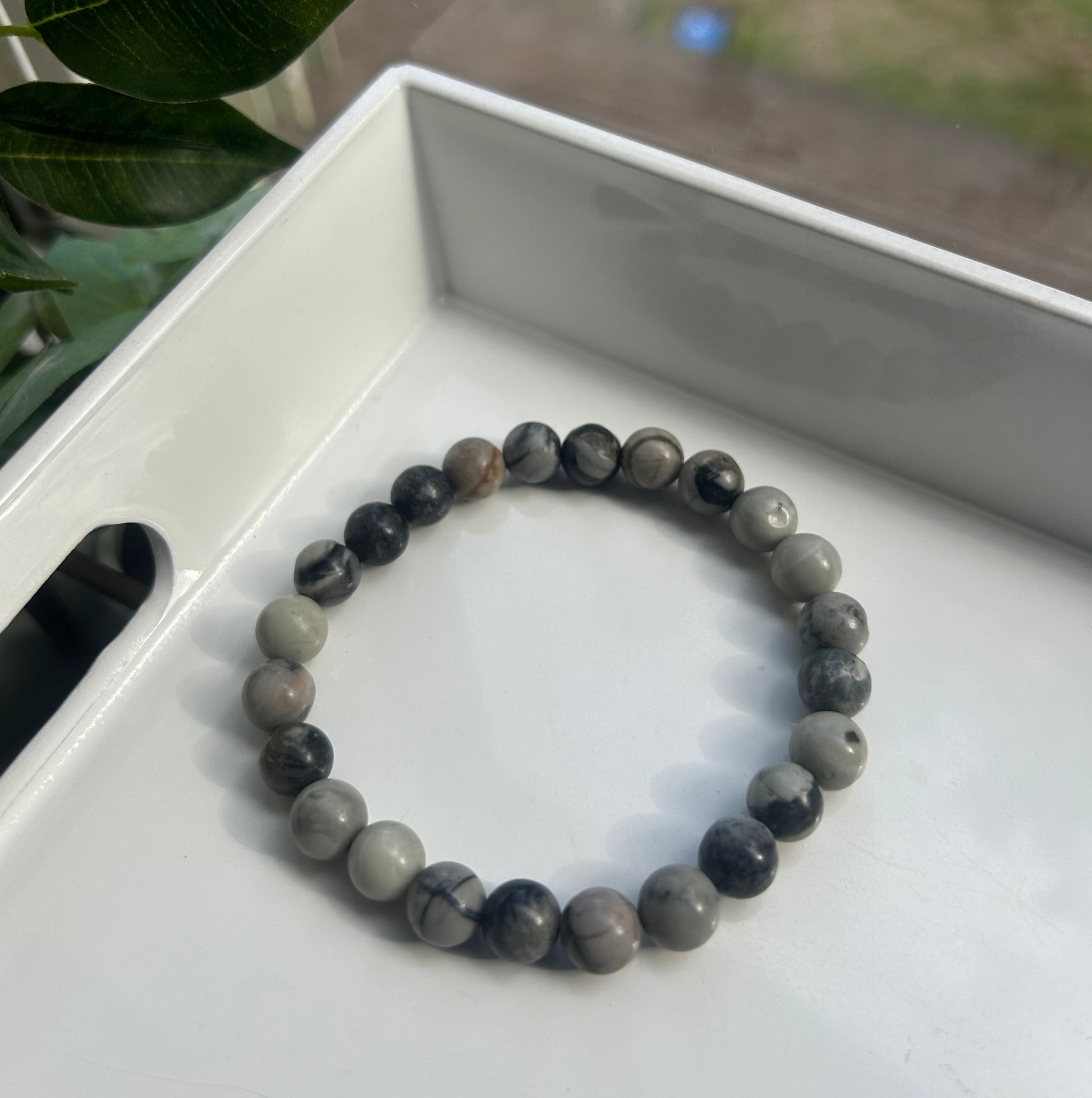 A beaded stretch bracelet made of round gray and black marbled gemstone beads, displayed inside a white tray with green leaves in the foreground.