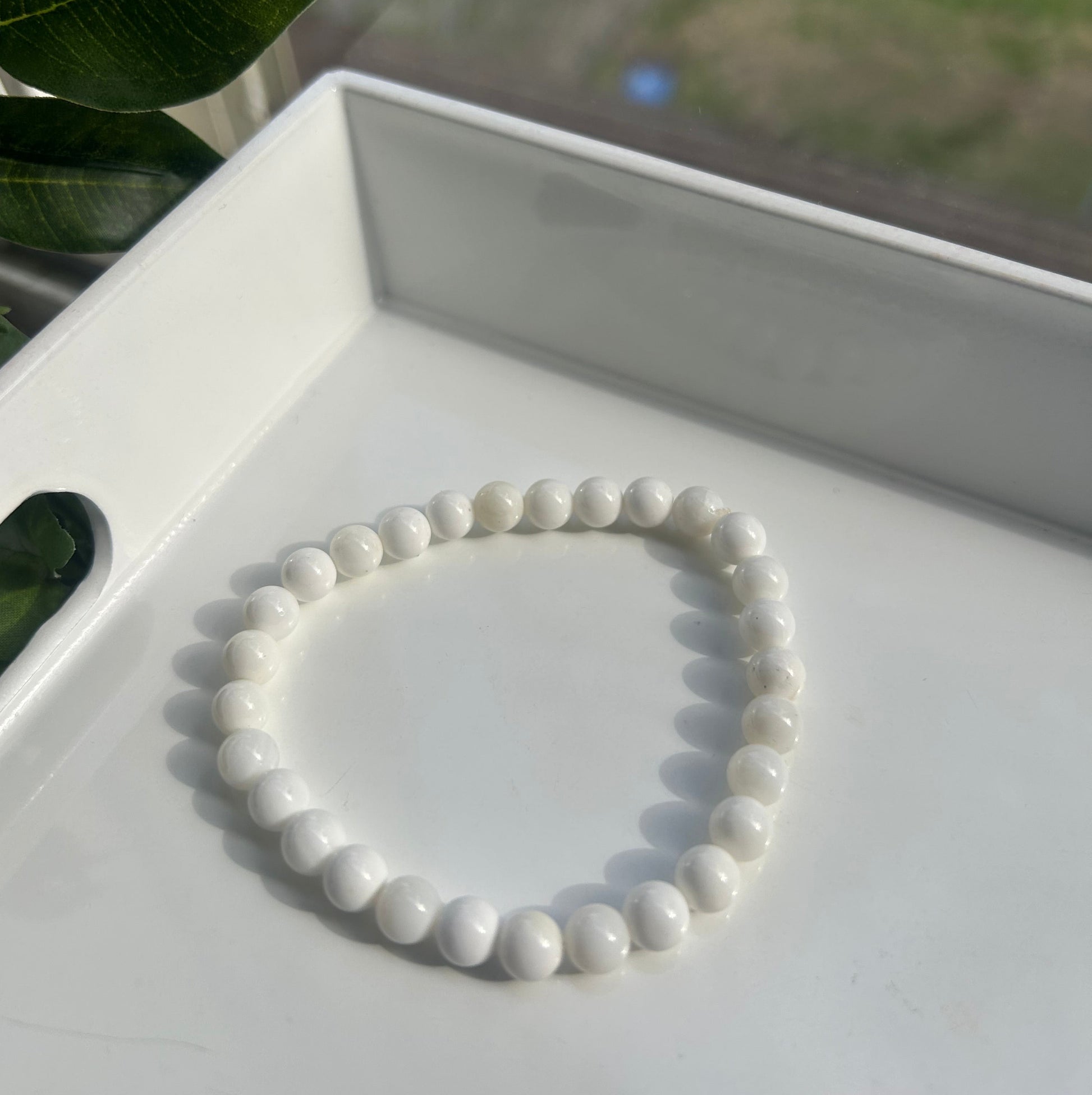 A white beaded bracelet made of smooth, glossy oval beads, displayed inside a white geometric tray with blurred green leaves in the foreground.