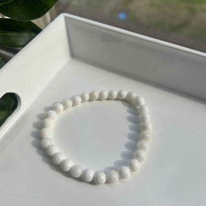 A white beaded bracelet made of smooth, glossy oval beads, displayed inside a white geometric tray with blurred green leaves in the foreground.