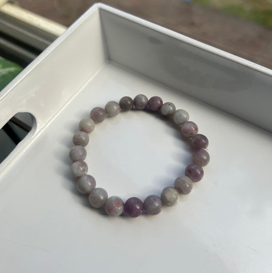 A beaded stretch bracelet made of round, polished beads in shades of purple, gray, and white, resting inside a white tray.