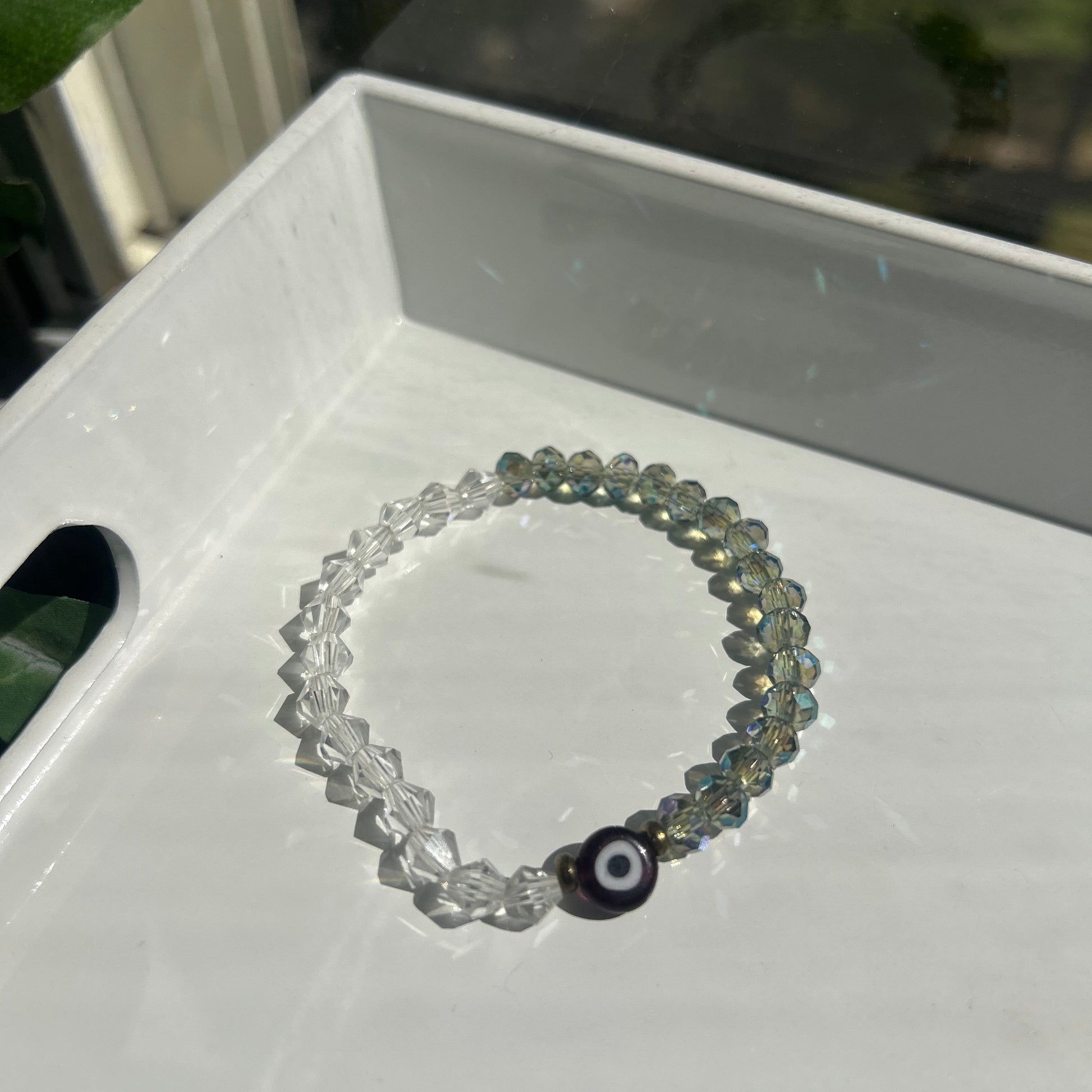 A beaded stretch bracelet with iridescent gray-green beads, clear faceted beads, and a black evil eye charm, resting on a white tray.