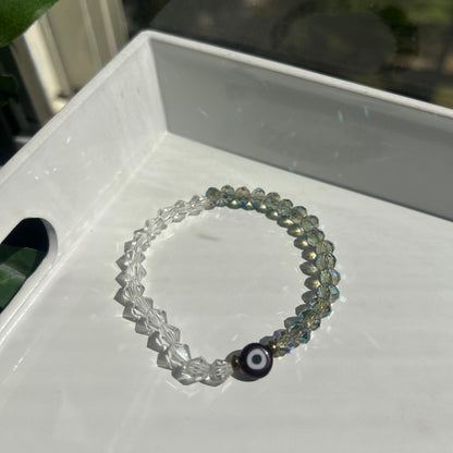 A beaded stretch bracelet with iridescent gray-green beads, clear faceted beads, and a black evil eye charm, resting on a white tray.