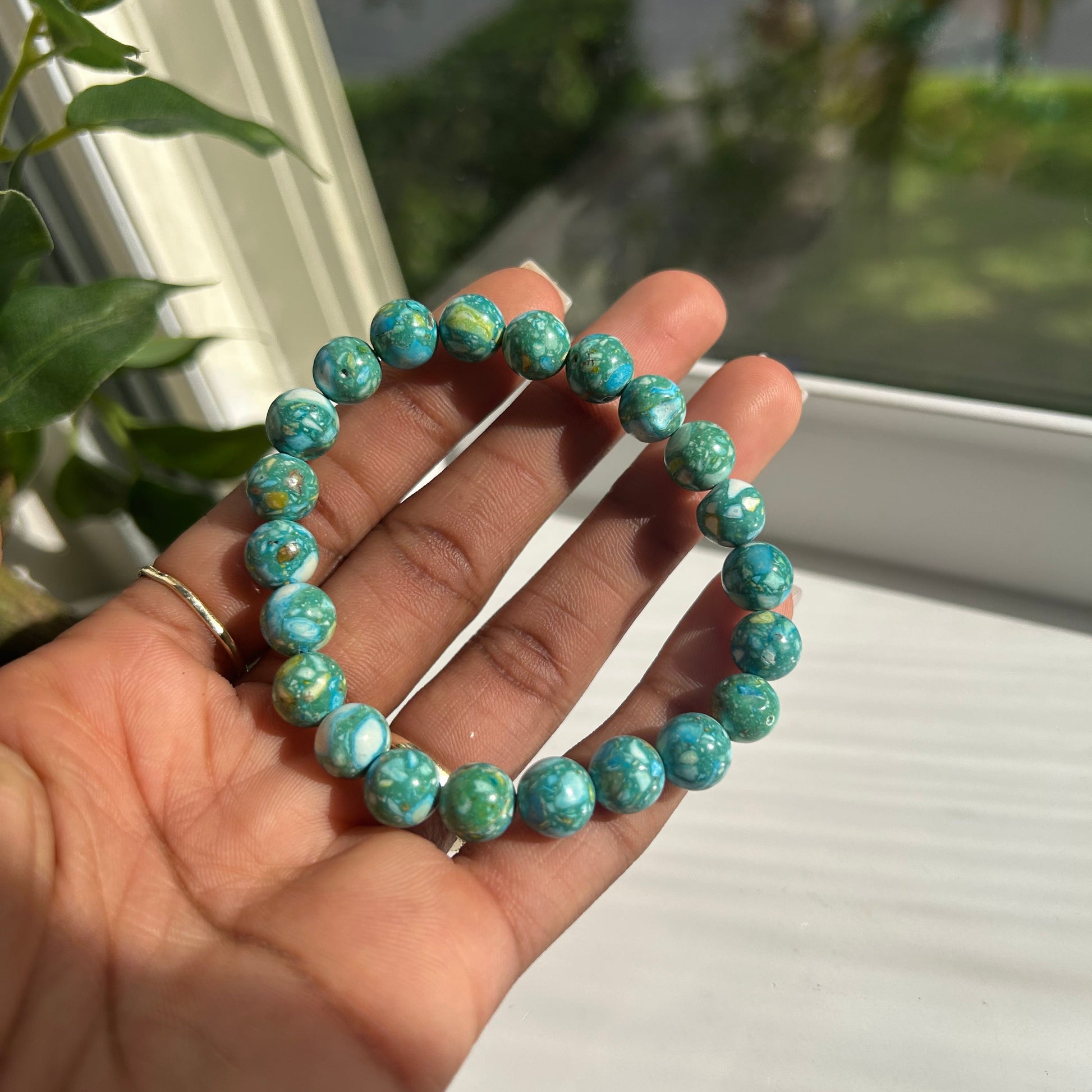 A beaded stretch bracelet made of turquoise-blue marbled beads, held in a person's hand near a window.