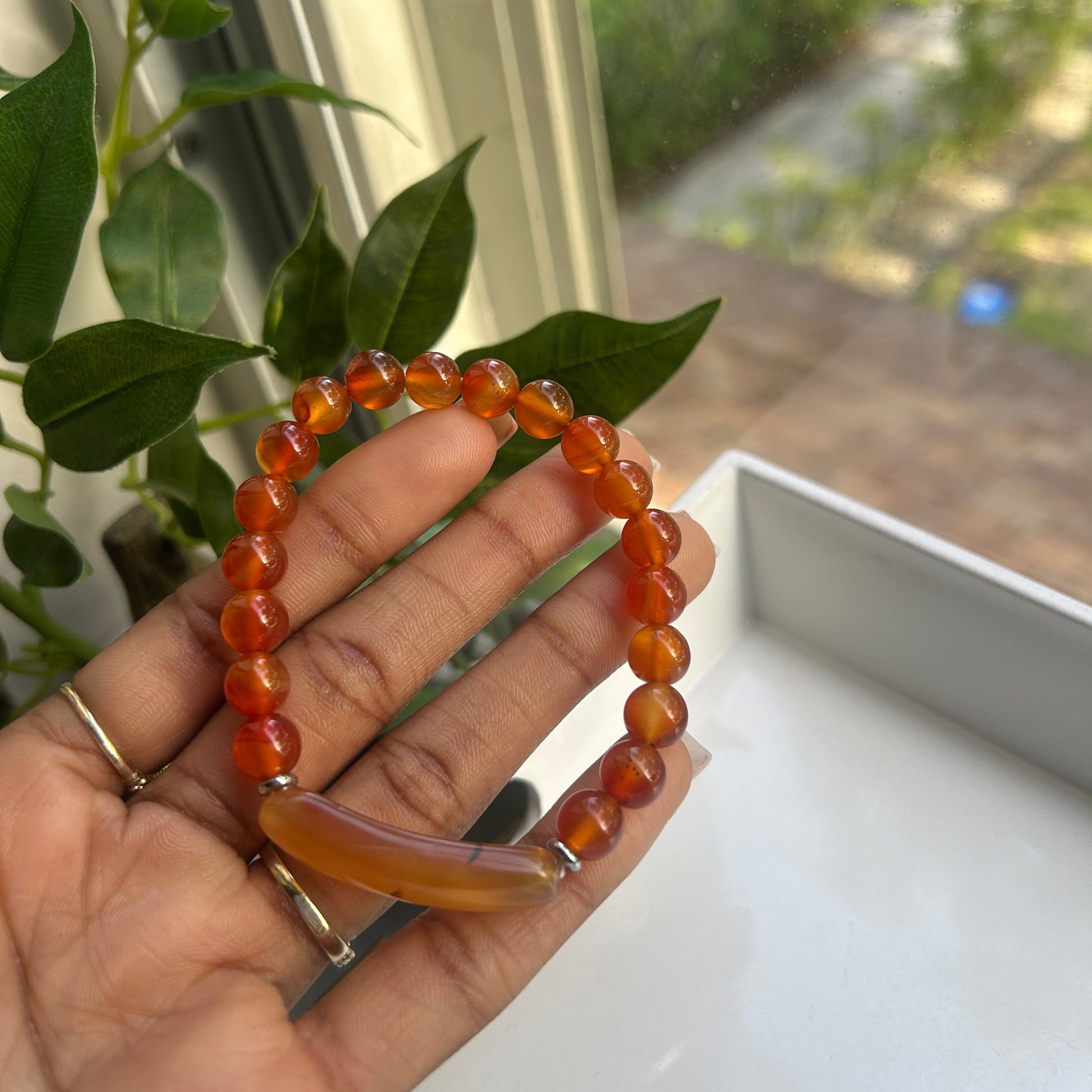A beaded stretch bracelet made of round orange-brown beads with a smooth, polished crescent moon pendant, held in a person's hand near a window with green leaves in the foreground.