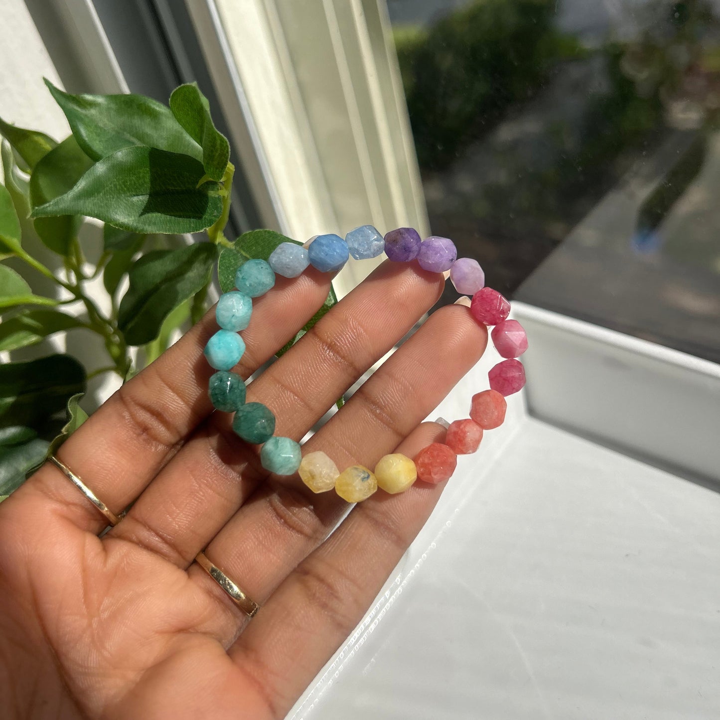 A beaded stretch bracelet with rainbow-colored round beads held in a person's hand, with a window and green plant leaves in the background.