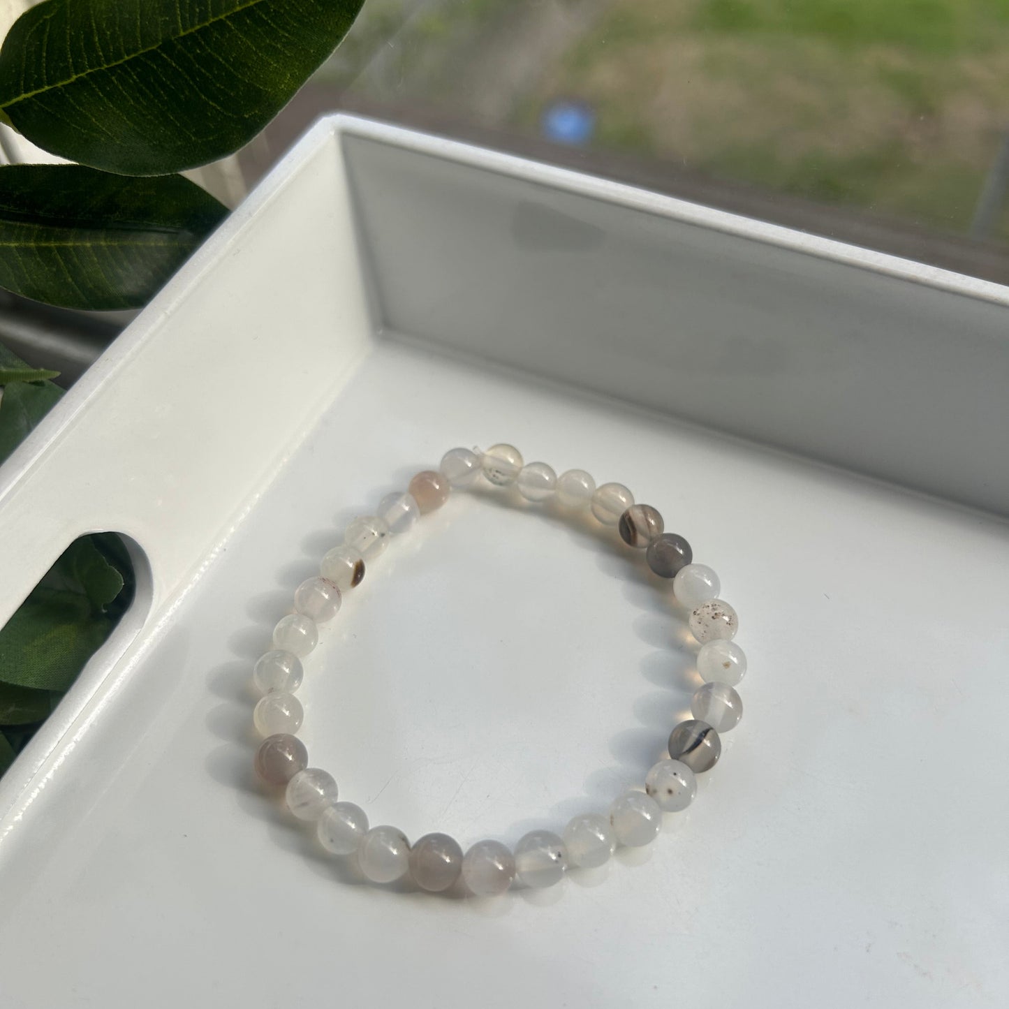 A beaded stretch bracelet made of translucent white and light gray round beads, resting inside a white tray with green leaves in the foreground.