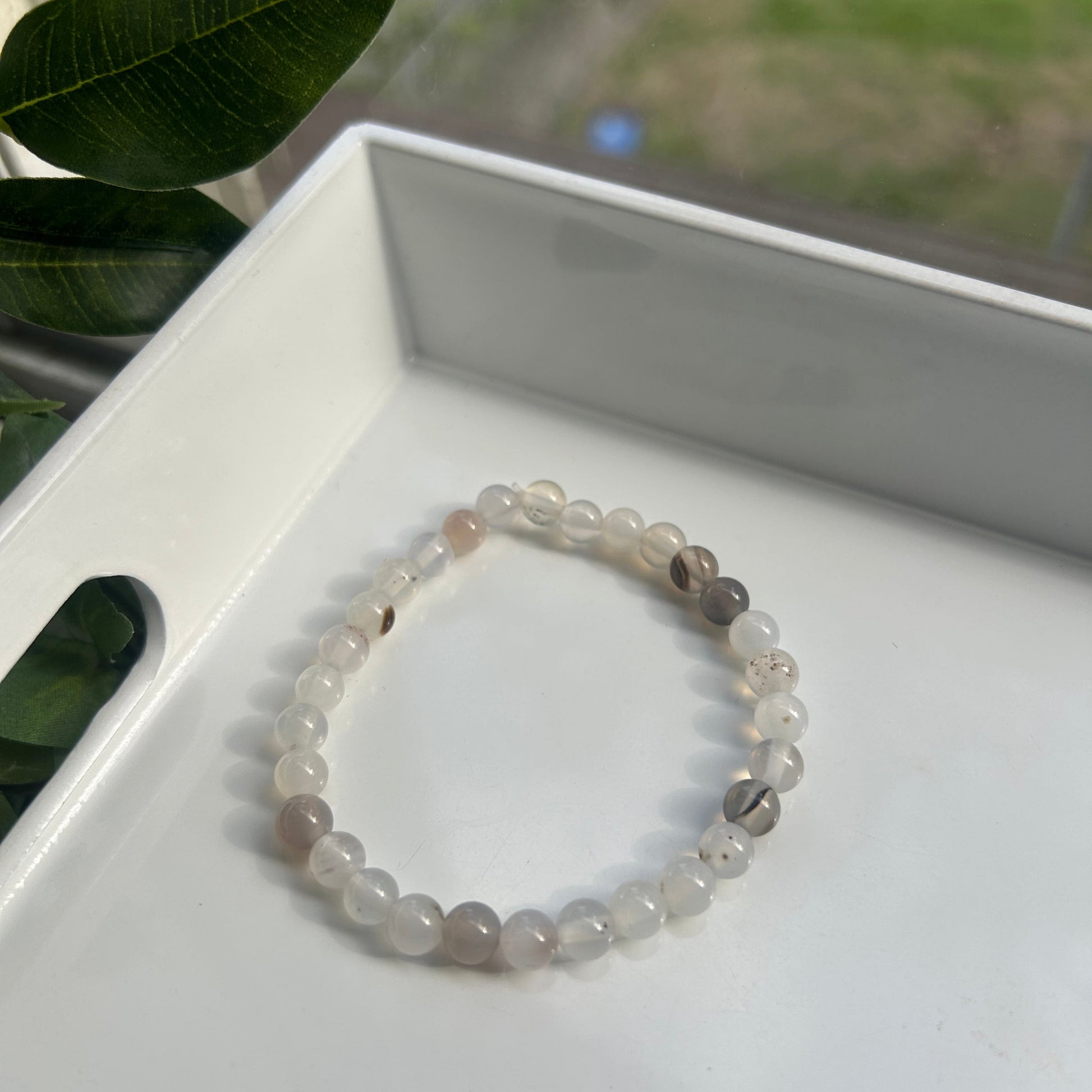 A beaded stretch bracelet made of translucent white and light gray round beads, resting inside a white tray with green leaves in the foreground.