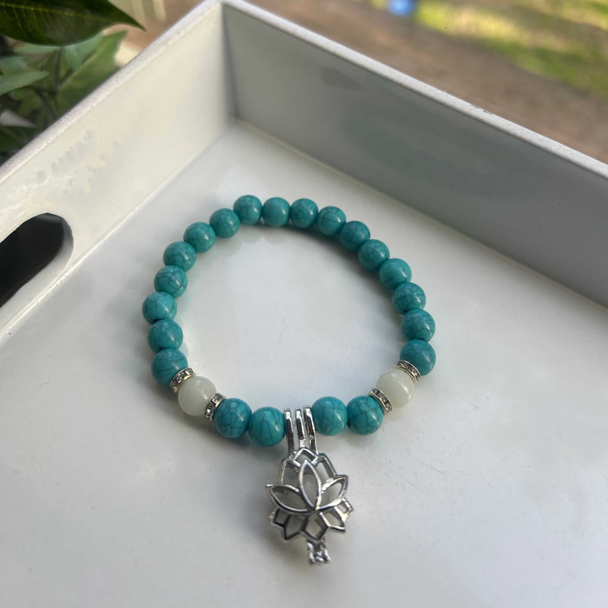 A beaded stretch bracelet with turquoise-blue round beads, white spacer beads, and a silver-toned lotus flower charm, displayed on a white tray.