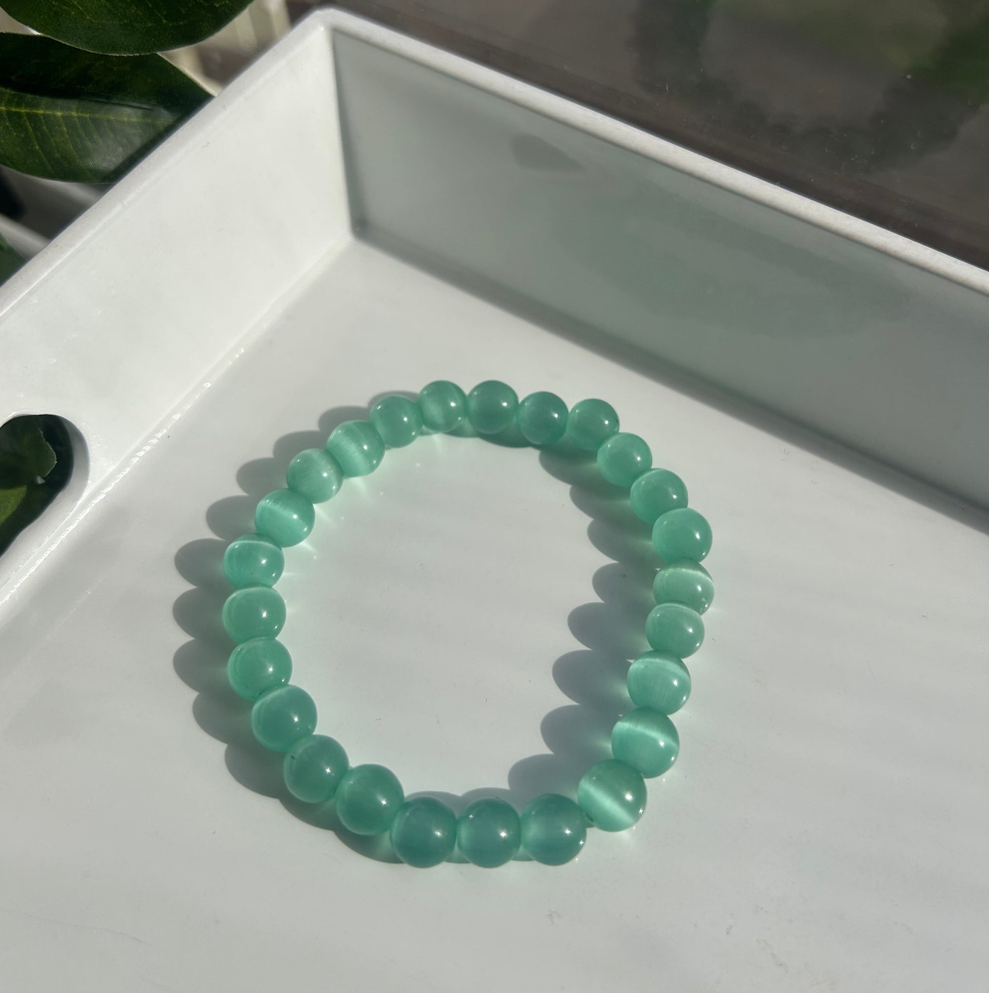 A beaded stretch bracelet made of translucent light green cat's eye beads, resting on a white tray with a leaf in the foreground.