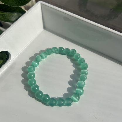 A beaded stretch bracelet made of translucent light green cat's eye beads, resting on a white tray with a leaf in the foreground.