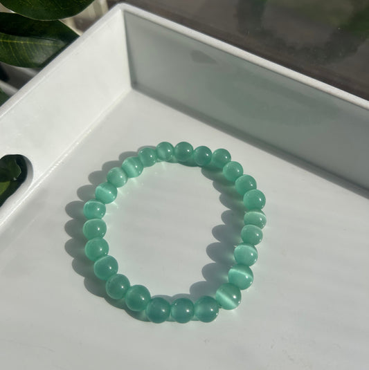 A beaded stretch bracelet made of translucent light green cat's eye beads, resting on a white tray with a leaf in the foreground.
