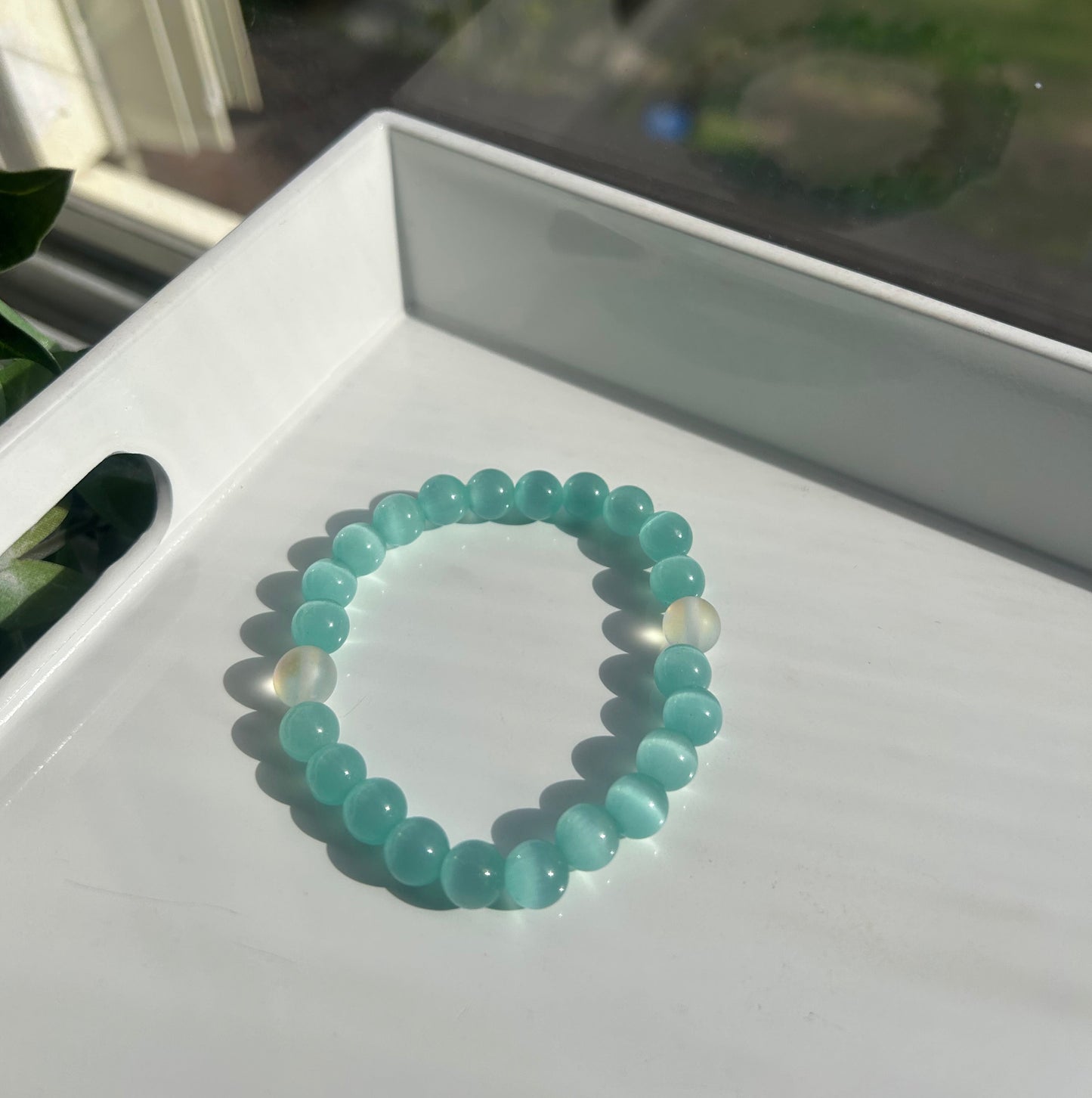 A beaded stretch bracelet made of light blue-green round beads with one translucent white bead, resting on a white tray.